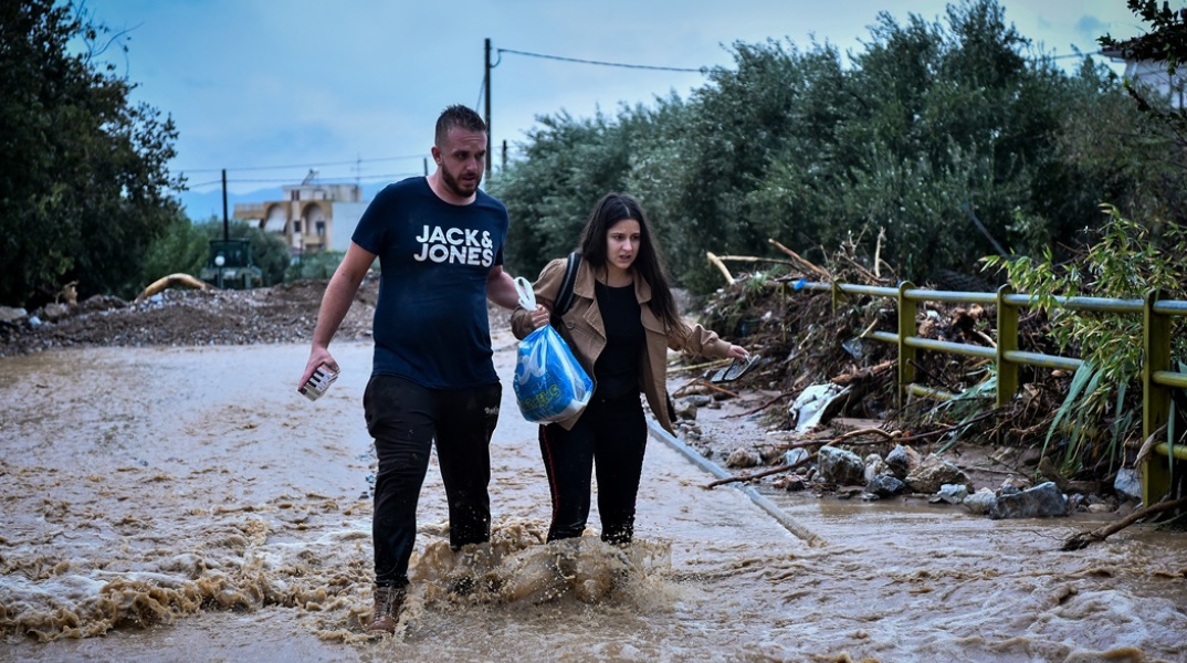 Γυναίκα και άνδρας βοηθούν ο ένας τον άλλο να περπατήσουν σε πλημμυρισμένο δρόμο στην Αγριά της Μαγνησίας
