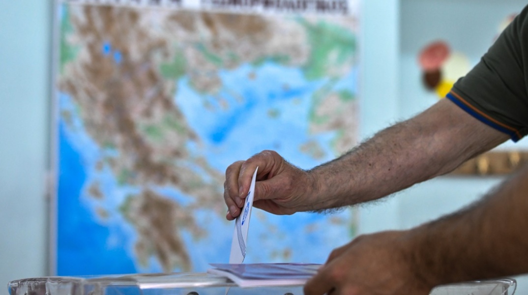 Χέρι ψηφοφόρου ρίχνει στην κάλπη το ψηφοδέλτιο - Πίσω του ο χάρτης της Ελλάδας
