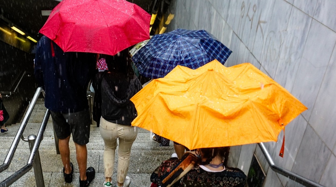 Πολίτες με ομπρέλες εν μέσω βροχής κατεβαίνουν τα σκαλιά προς την είσοδο του μετρό 