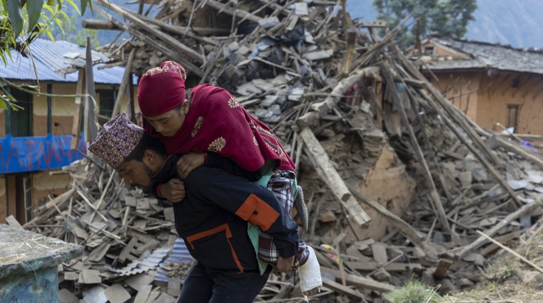 Άνδρας μεταφέρει γυναίκα στην πλάτη του ενώ πίσω τους υπάρχουν χαλάσματα από τον σεισμό στο Νεπάλ