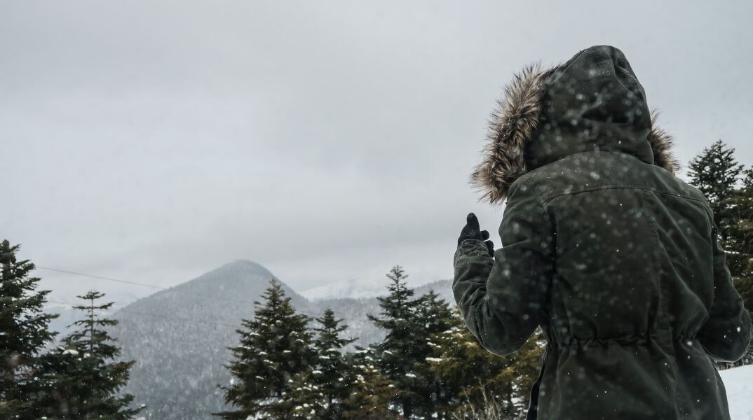 Γυναίκα με μπουφάν και κουκούλα απολαμβάνει τα χιόνια σε χιονισμένο τοπίο με δέντρα 
