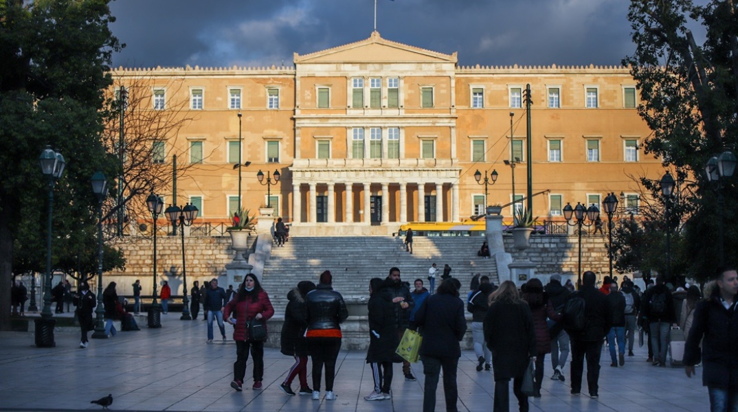 Πολίτες περπατούν στην πλατεία Συντάγματος με φόντο τη Βουλή των Ελλήνων