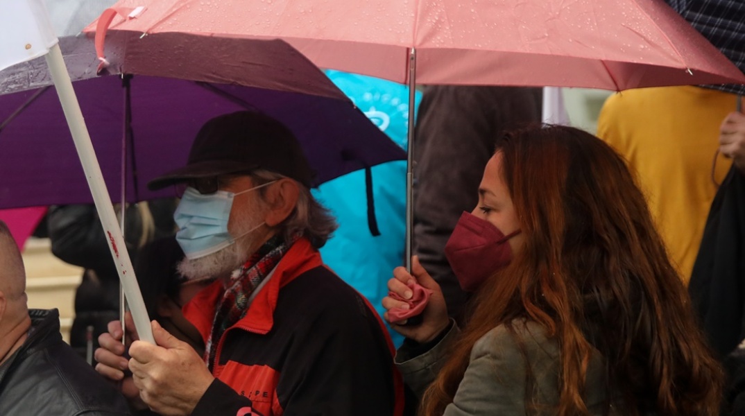 Γυναίκα με ομπρέλα φορά μάσκα δίπλα σε άνδρα επίσης με μάσκα και ομπρέλα