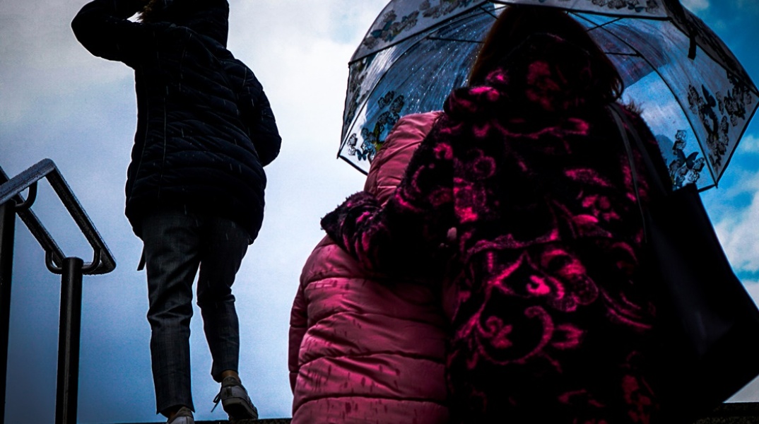 Γυναίκα με ομπρέλα κρατά κοντά της το παιδί της για να μην βραχεί όσο ανεβαίνουν σκάλες