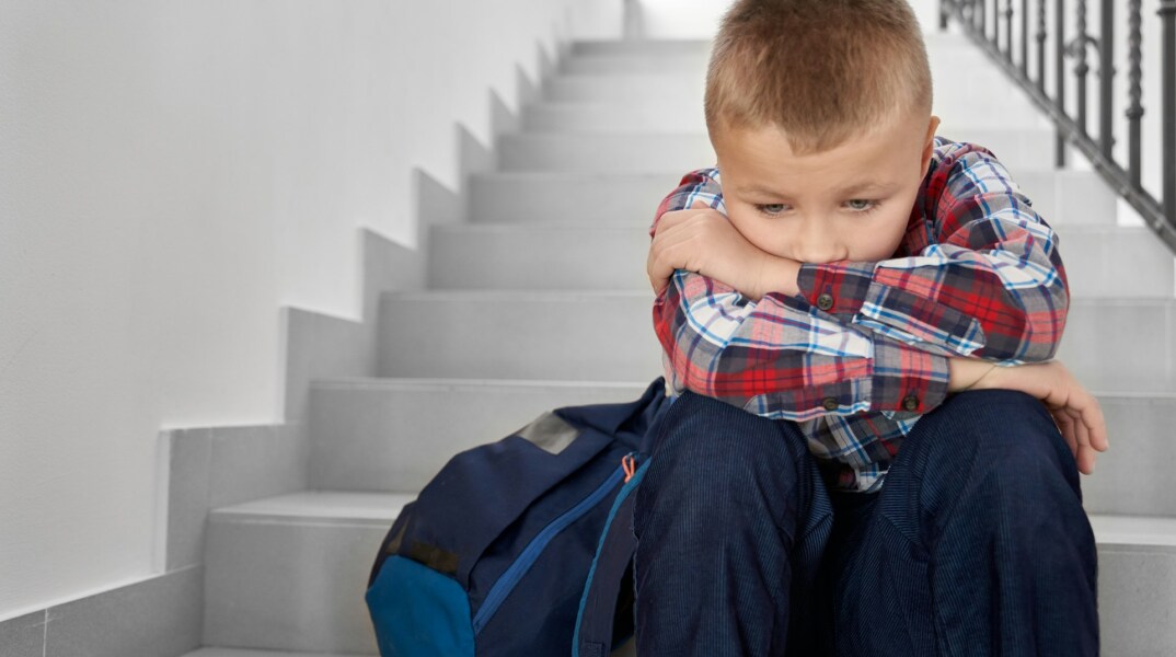 Παιδί κάθεται στα σκαλιά με την τσάντα του δίπλα και έχει ακουμπήσει το κεφάλι του στα γόνατά του - Εικόνα που παραπέμπει σε βία στα σχολεία - bullying