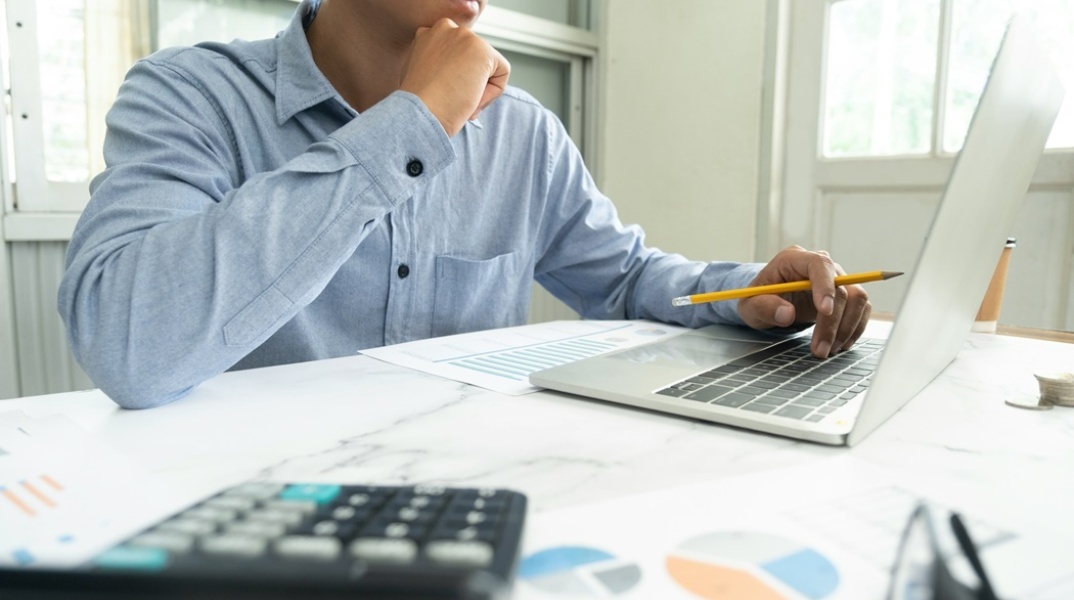 Άτομο μπροστά από λάπτοπ με χαρτιά που απεικονίζουν γραφήματα κάνει υπολογισμούς