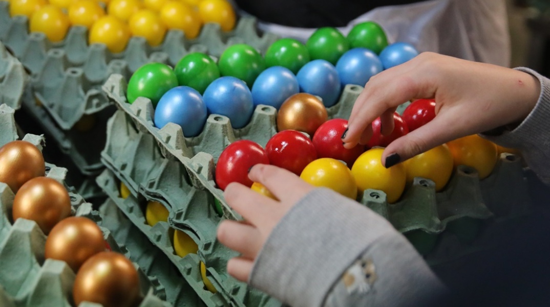 Αυγά βαμμένα σε μπλε, κόκκινο, κίτρινο και πράσινο χρώμα μέσα σε αυγοθήκες και ένα χέρι που επιλέγει ποια θα αγοράσει