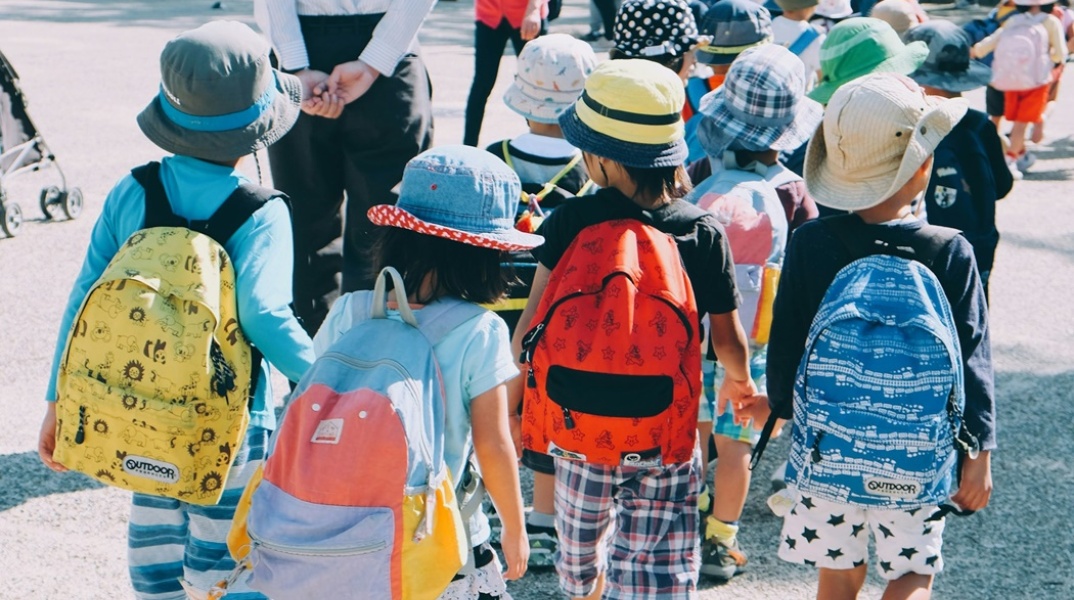 Παιδιά με καλοκαιρινά ρούχα και τσάντες στην πλάτη περπατούν πιασμένα χεράκι - χεράκι