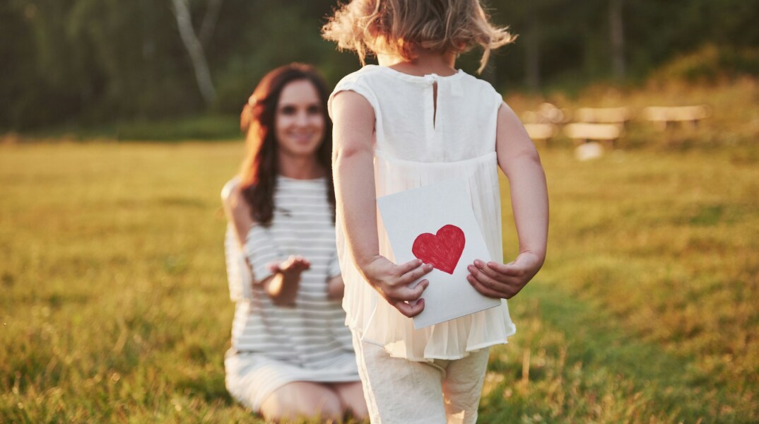 Κοριτσάκι κρύβει στην πλάτη της μία καρδούλα όσο πλησιάζει προς τη μητέρα της που έχει ανοίξει την αγκαλιά της για να τη δεχθεί