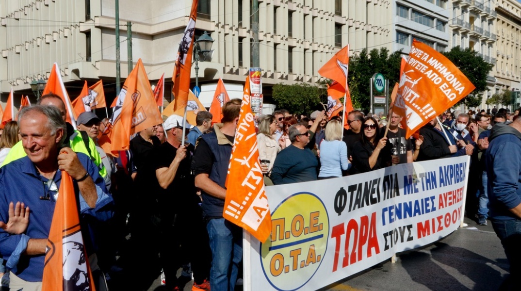 Άτομα που συμμετέχουν σε συγκέντρωση στο κέντρο της Αθήνας κρατώντας σημαίες και πανό της ΠΟΕ-ΟΤΑ