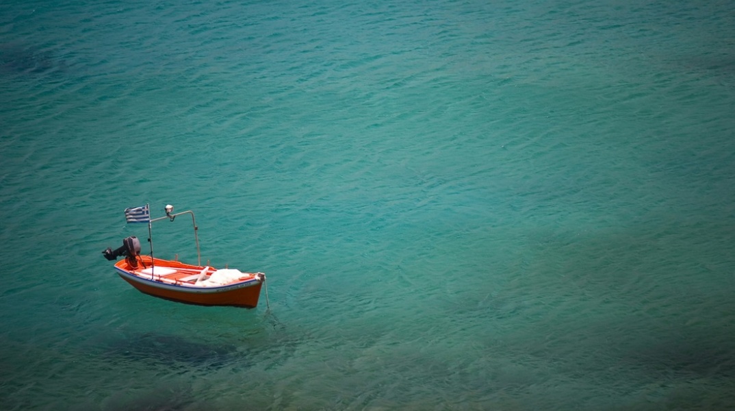 Παραλία και καΐκι αγκυροβολημένο που έχει στην πλώρη του την ελληνική σημαία