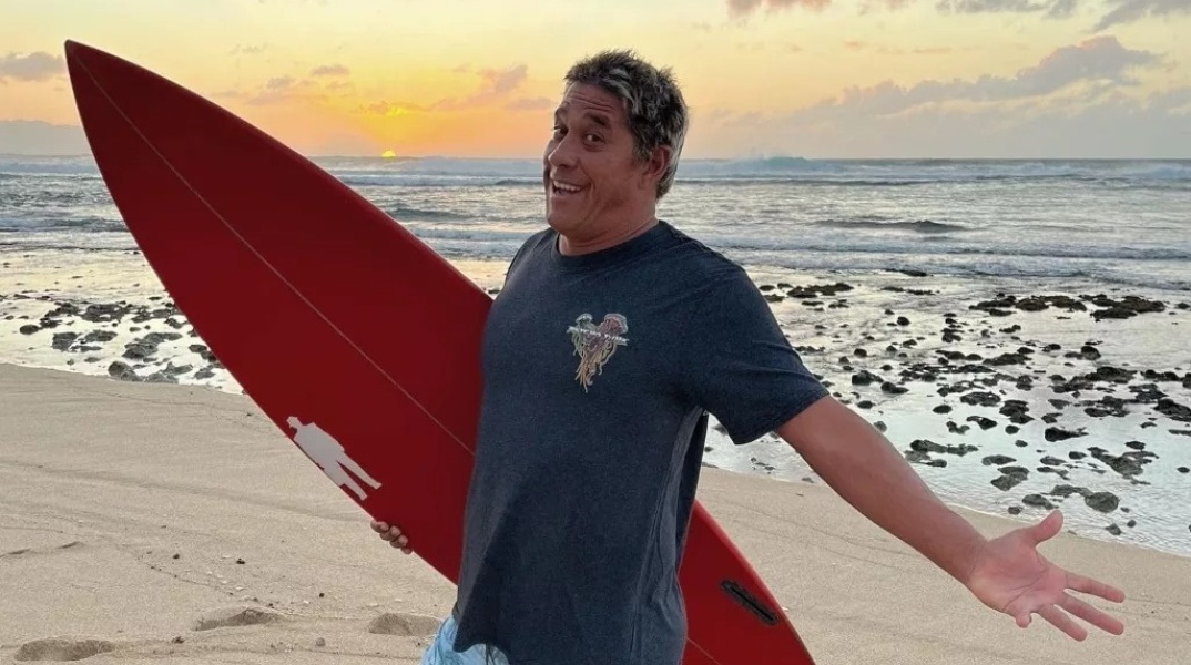 Ο Ταμάγιο Πέρι ποζάρει σε παραλία με τη σανίδα του σερφ ανά χείρας