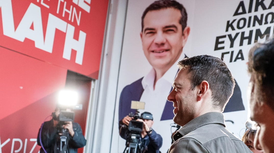 Ο Στέφανος Κασσελάκης προχωρά σε κτίριο που στο εξωτερικό του έχει αφίσα του ΑΛέξη Τσίπρα