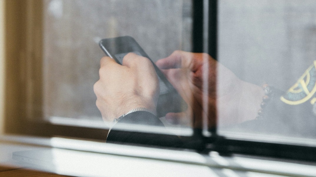 Άνδρας πίσω από τζάμι κρατά και πληκτρολογεί στο κινητό του τηλέφωνο