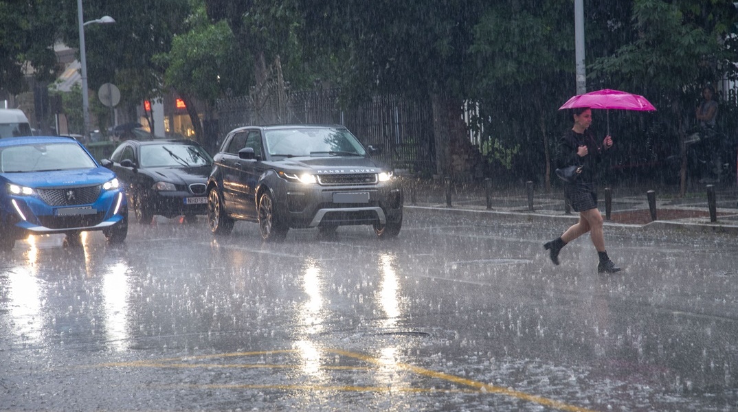 Γυναίκα με ομπρέλα διασχίζει δρόμο υπό καταρρακτώδη βροχή