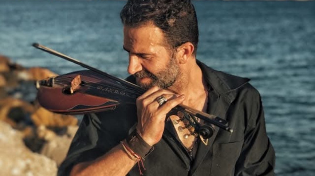 Ο Ηλίας Παλιουδάκης μπροστά από τη θάλασσα κρατώντας παραδοσιακό μουσικό όργανο