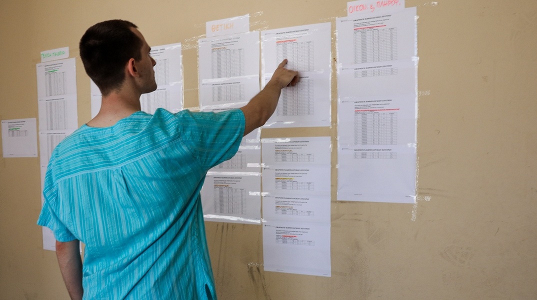 Άτομο μπροστά από τις βαθμολογίες των μαθημάτων των Πανελληνίων που είναι ανηρτημένες έξω από σχολείο