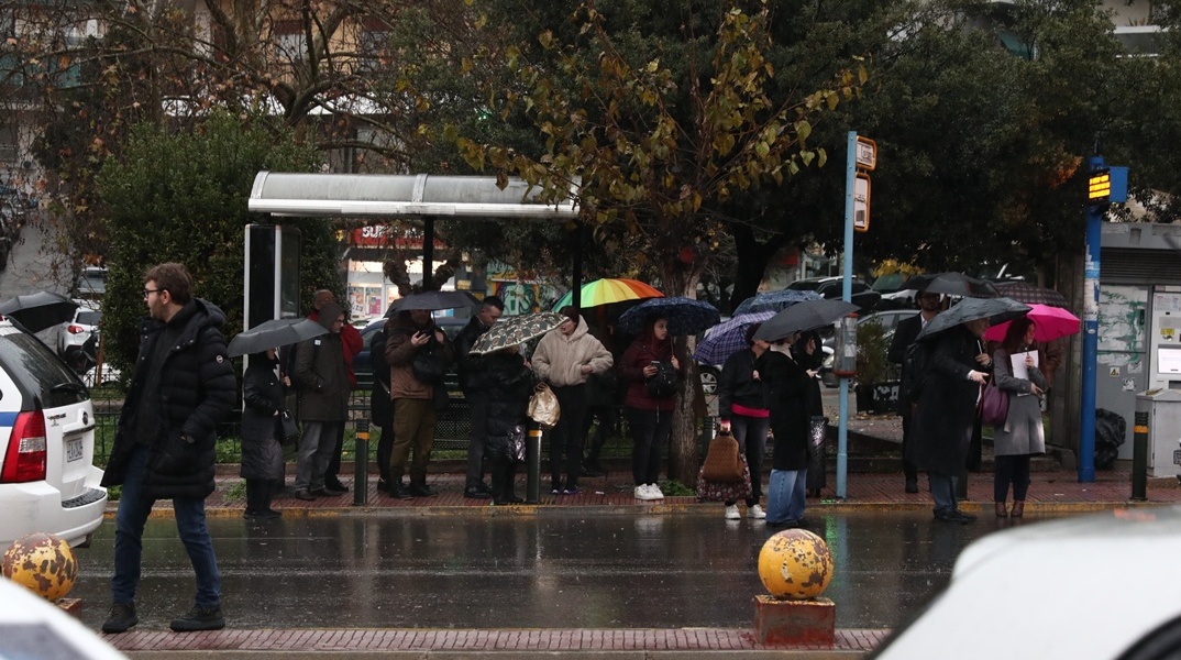 Πολίτες σε στάση λεωφορείου περιμένουν έχοντας ανοιχτή την ομπρέλα τους