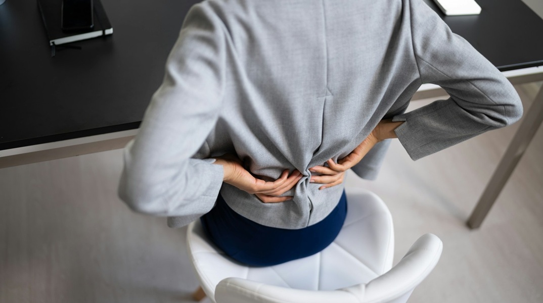 Εργαζόμενη κάθεται σε καρέκλα γραφείου και κρατά τη μέση της που φαίνεται ότι πονά