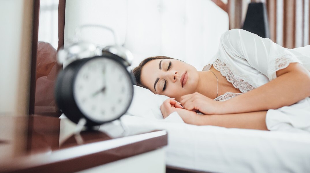Γυναίκα κοιμάται στο κρεβάτι έχοντας στο κομοδίνο ένα ξυπνητήρι