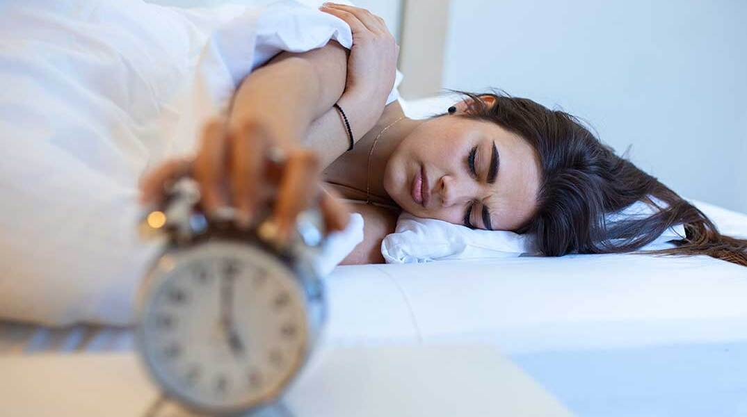 Γυναίκα κοιμάται στο κρεβάτι με φόντο το ξυπνητήρι