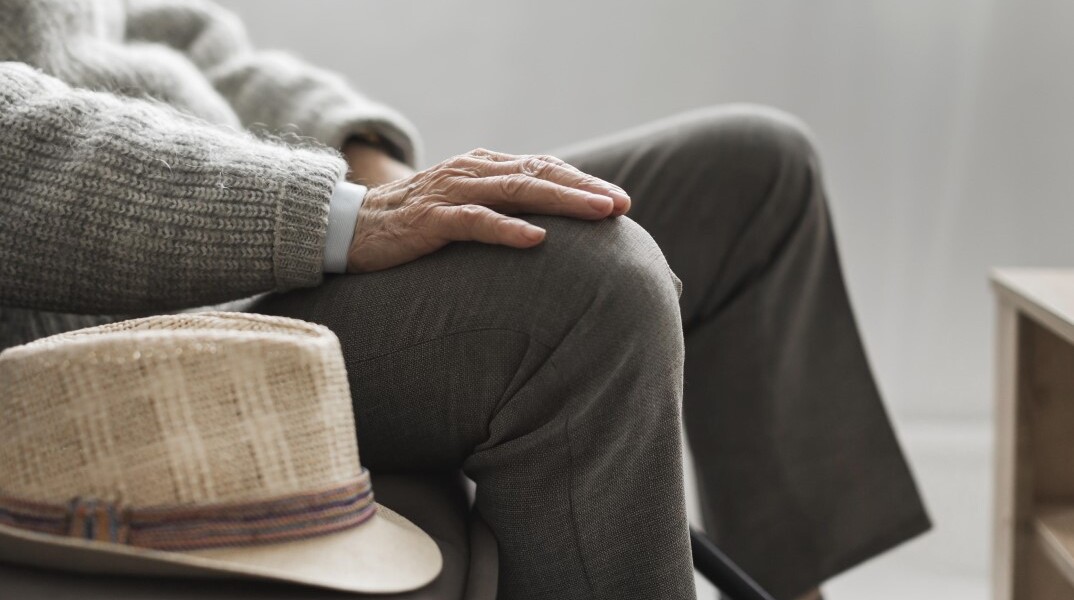 Ηλικιωμένος με καπέλο ακουμπισμένο στο πόδι κάθεται σε καναπέ