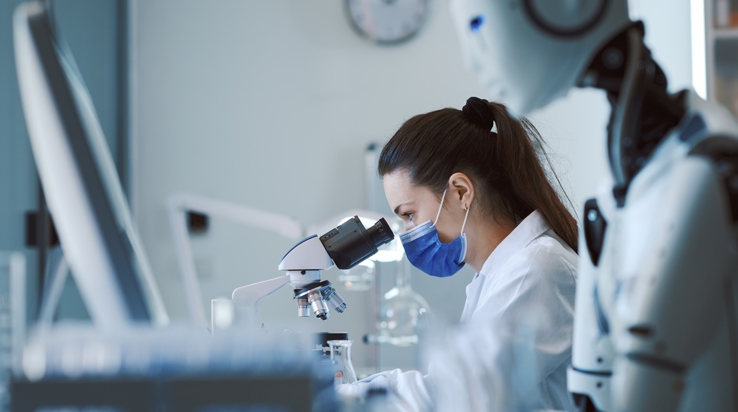 Γυναίκα με μάσκα που κοιτά σε μικροσκόπιο και δίπλα της ένα ρομπότ