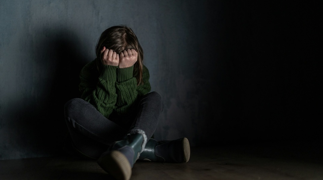 Ανήλικη που κρύβει το πρόσωπό της με τα χέρια της καθισμένη σε ένα σκοτεινό δωμάτιο