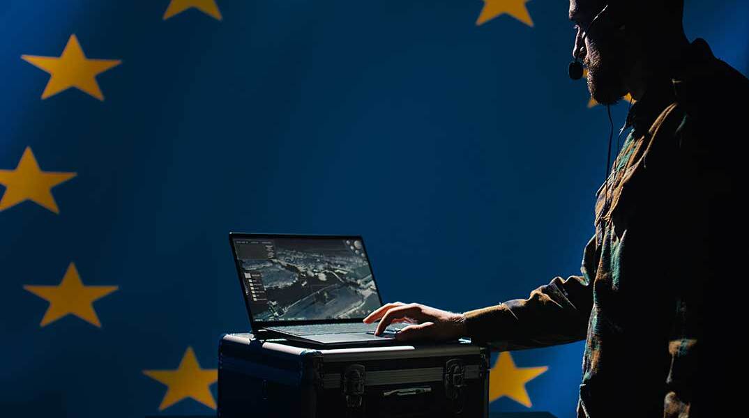 Στρατιωτικός αναλυτής μπροστά σε υπολογιστή με φόντο τη σημαία της ΕΕ
