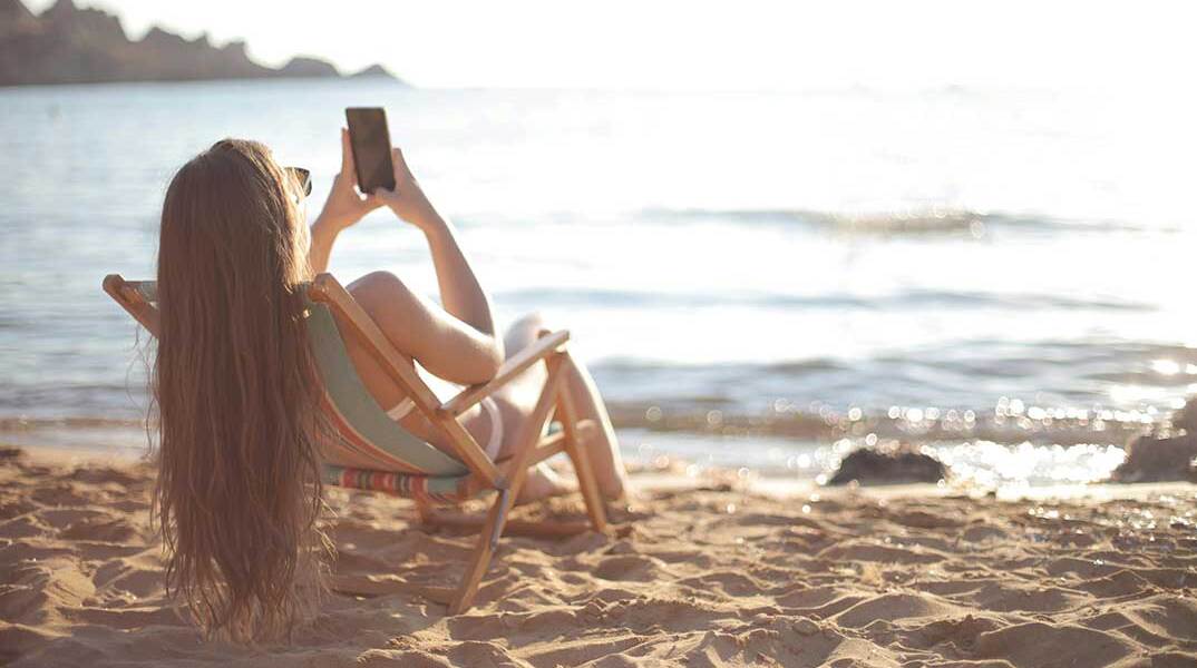 Γυναίκα σε ξύλινη καρέκλα στην παραλία κοιτά το κινητό της τηλέφωνο