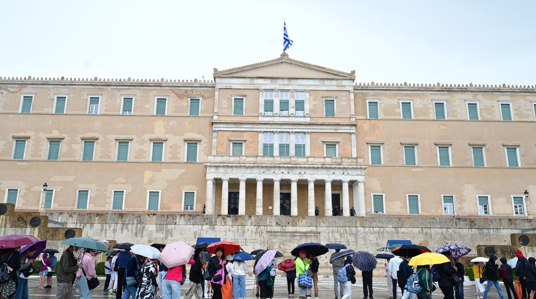 Πολίτες με ομπρέλες εν μέσω βροχής μπροστά από το μνημείο του Άγνωστου Στρατιώτη και τη Βουλή