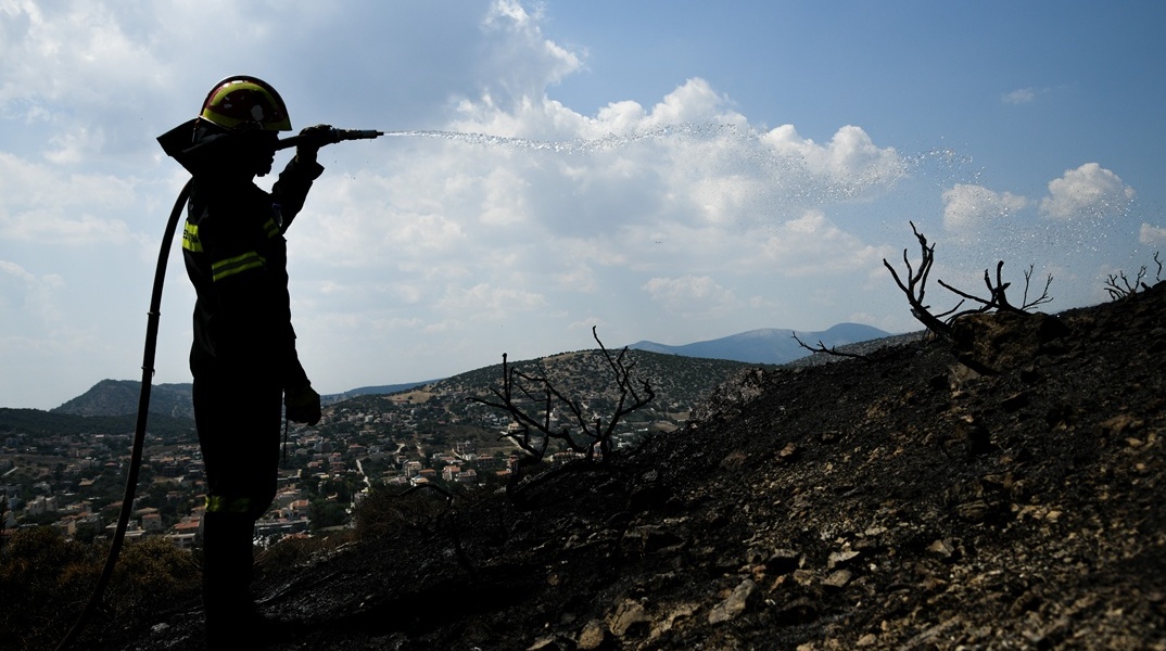 Πυροσβέστης με τη μάνικα ανά χείρας ρίχνει νερό σε καμένο τοπίο