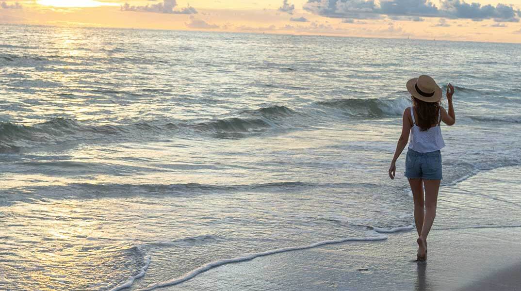 Γυναίκα με ψάθινο καπέλο και τζιν σορτσάκι στην παραλία με φόντο το ηλιοβασίλεμα