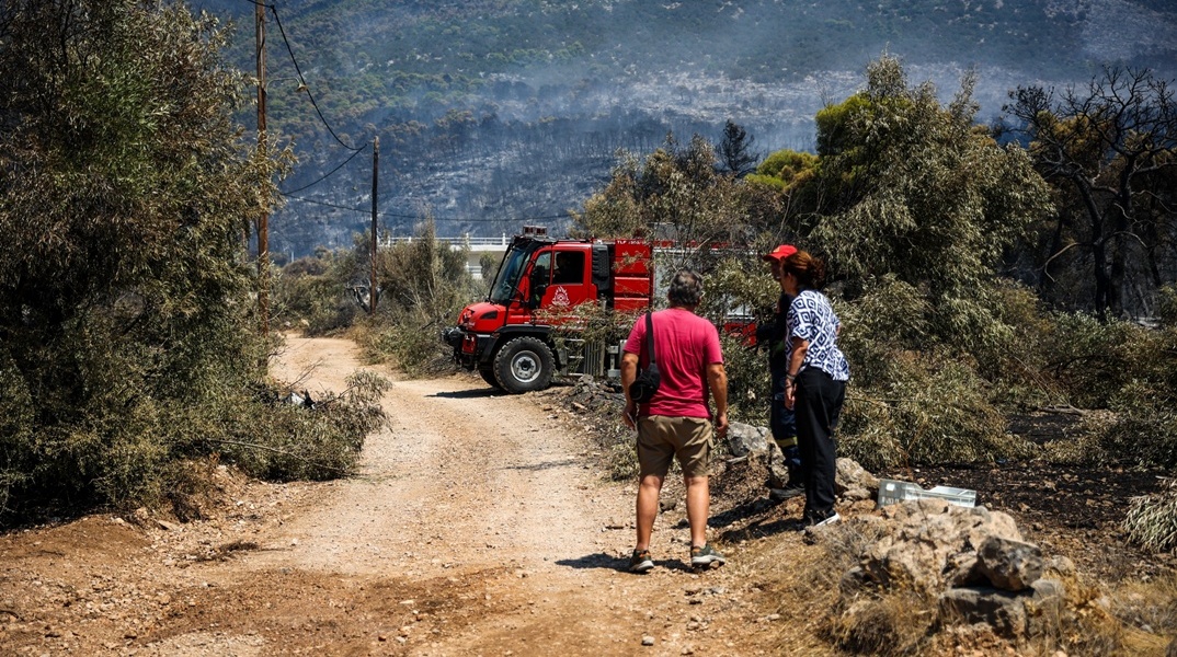Πυροσβεστικό όχημα και πολίτες δίπλα σε αυτό με φόντο καμένη έκταση στο βάθος της φωτογραφίας