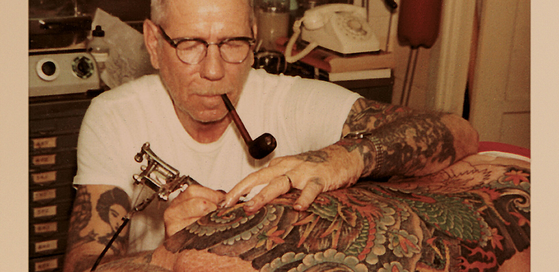 Norman Collins tattooing back with pipe in mouth