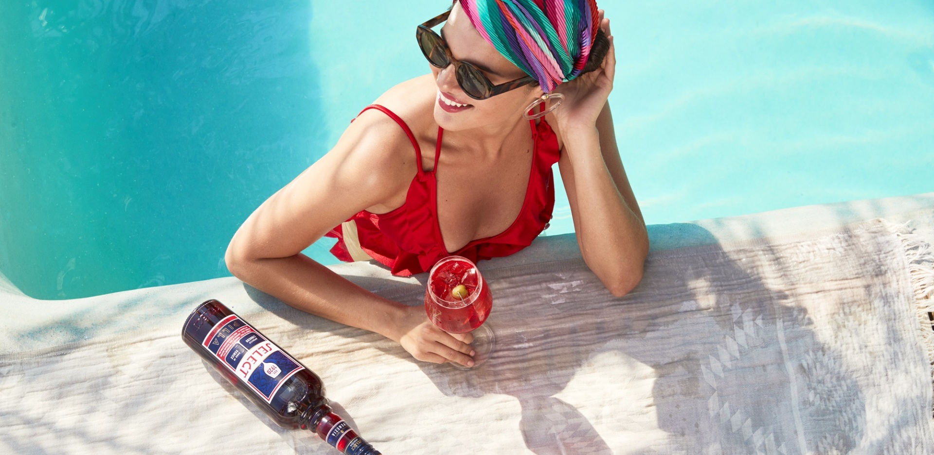 Γυναίκα σε πισίνα κρατάει ποτήρι με Spritz Veneziano και δίπλα είναι το μπουκάλι Select Aperitivo