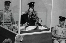 Ο Ναζί εγκληματίας πολέμου Άντολφ Άιχμαν στέκεται σε προστατευτικό γυάλινο θάλαμο πλαισιωμένο από την ισραηλινή αστυνομία κατά τη διάρκεια της δίκης του στις 22 Ιουνίου 1961 στην Ιερουσαλήμ