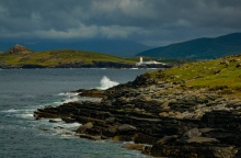 Βραχώδης παραλία και στο βάθος άσπρο κτίριο και κόκκινος φάρος