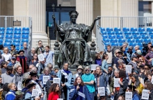 Καθηγητές ντυμένοι με παραδοσιακές ρόμπες μπροστά από άγαλμα κρατούν πλακάτ