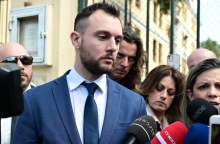 Άνδρας με κοστούμι μπροστά σε μικρόφωνα ανάμεσα από δημοσιογράφους