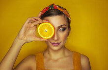 Καστανή κοπέλα με μαζεμένα μαλλιά κρατάει μια φέτα πορτοκάλι μπροστά από το αριστερό της μάτι, σε πορτοκαλί φόντο