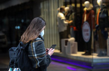 Κοπέλα με μάσκα για τον κορωνοϊό κοιτάζει το κινητό της με φόντο καταστήματα στην Ερμού