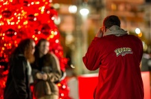 Άνδρας ντυμένος στα κόκκινο βγάζει φωτογραφία δύο κοπέλες μπροστά από ένα κόκκινο χριστουγεννιάτικο δέντρο