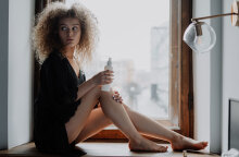 Κοπέλα με σγουρά μαλλιά καθισμένη δίπλα σε παράθυρο φορώντας μαύρα ρούχα βάζει κρέμα στα πόδια της