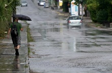 Πολίτης με ομπρέλα σε πεζοδρόμιο και δίπλα του αυτοκίνητο διασχίζει πλημμυρισμένο δρόμο