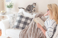 Γυναίκα καθισμένη σε καναπέ διαβάζει βιβλίο, δίπλα κάθεται γατάκι