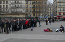 Άνθρωποι που περιμένουν σε μια ουρά και μια άστεγη γυναίκα κάθεται κοντά τους στην πλατεία Madeleine στο Παρίσι