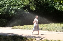 Γυναίκα περπατά σε συνθήκες καύσωνα σε περιβάλλον όπου ποτιστικό δροσίζει φυτά πετώντας νερό