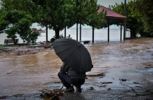 Άνδρας κρατά ομπρέλα για να προστατευτεί από καταιγίδα και στέκεται μπροστά από δρόμο που έχει μετατραπεί σε ποτάμι