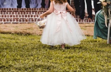 Κοριτσάκι με φόρεμα που παραπέμπει σε γάμο διασχίζει σημείο με γρασίδι και κρατά στα χέρια του καλαθάκι